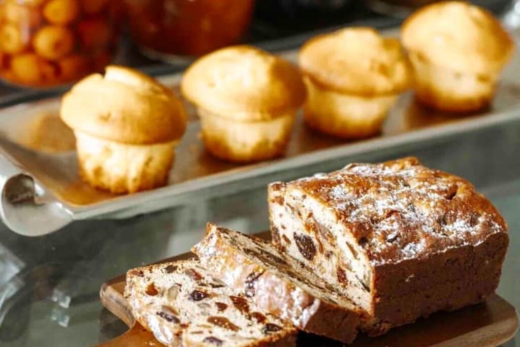 A loaf of bread with raisins on a wooden board, with several muffins in the background.