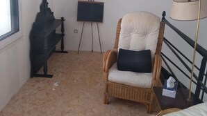 Une pièce avec un fauteuil en osier, une petite table avec une lampe, un tableau noir et une fenêtre.