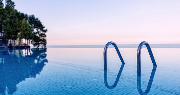 Una piscina infinita azul cristalina con dos escaleras metálicas, rodeada de árboles y un mar tranquilo al fondo.