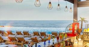 Un bar con vistas al océano, sillones de madera y diversas bebidas.