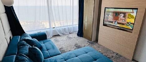 A modern living room with a blue sofa, a television on the wall, and a large window with sheer curtains.