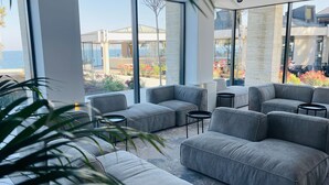 A modern outdoor seating area with grey sofas and round tables, overlooking a landscaped garden and a building with a glass facade.