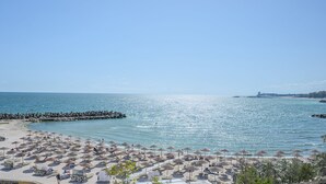 A beach with rows of sun umbrellas and lounge chairs, a rocky breakwater, and a clear blue sky.