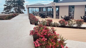A modern outdoor seating area with tables and chairs, surrounded by potted plants and a view of the sea.