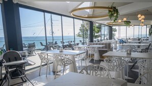 A modern outdoor dining area with white furniture, large windows offering ocean views, and hanging plants.