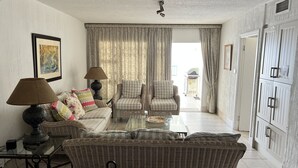 Sala de estar con un sofá, dos sillones, una mesa de centro y una lámpara. Se observa un ventanal con cortinas, una puerta con espejo y una pared decorada con un cuadro enmarcado.