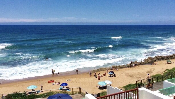 Una playa con gente y sombrillas, cielo despejado y olas rompiendo en la orilla.
