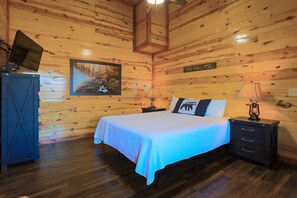 A wooden cabin bedroom with a bed, a nightstand, a lamp, a painting, and a sign on the wall. - Heated Pool|Cabin with Gorgeous Mountain Views (Sevierville)