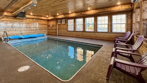 An indoor swimming pool with wooden walls and ceiling, a blue pool mat, and several Adirondack chairs.