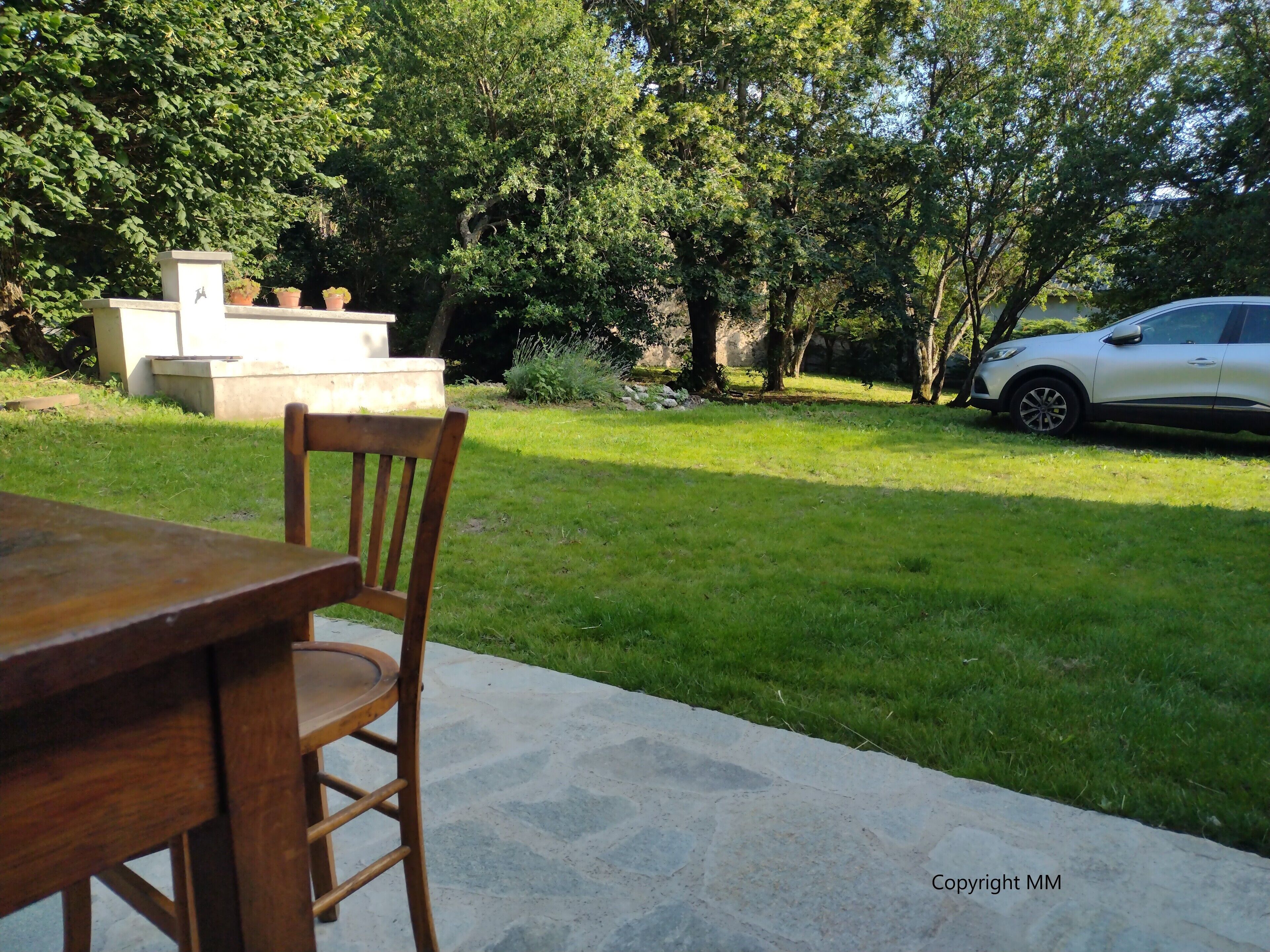 An outdoor patio with a wooden table and chair, a concrete bench, and a white car parked nearby.