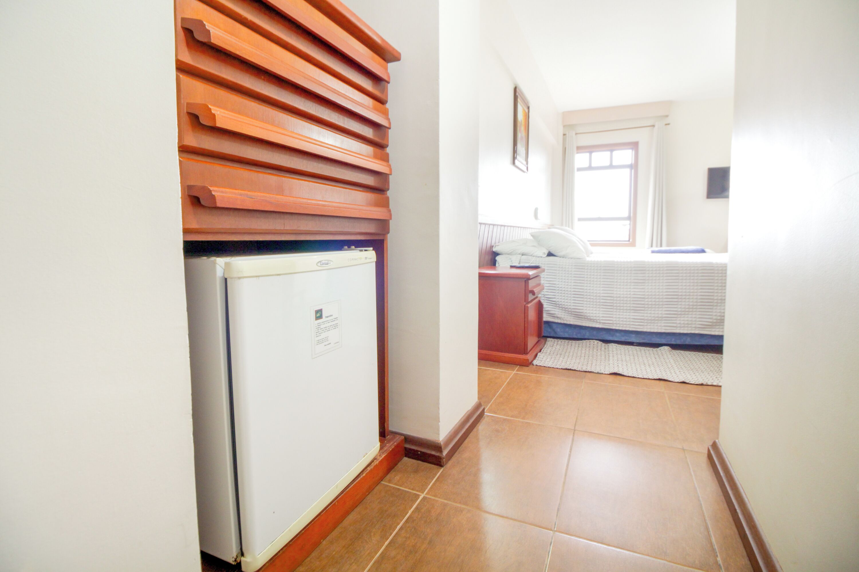 A narrow hallway with a white refrigerator, wooden slatted wall, and a bed with a blue mattress and white bedding.