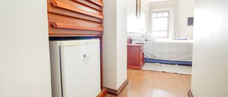 A narrow hallway with a white refrigerator, wooden slatted wall, and a bed with a blue mattress and white bedding.