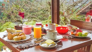 A breakfast table with various food items and drinks, set against a backdrop of greenery.