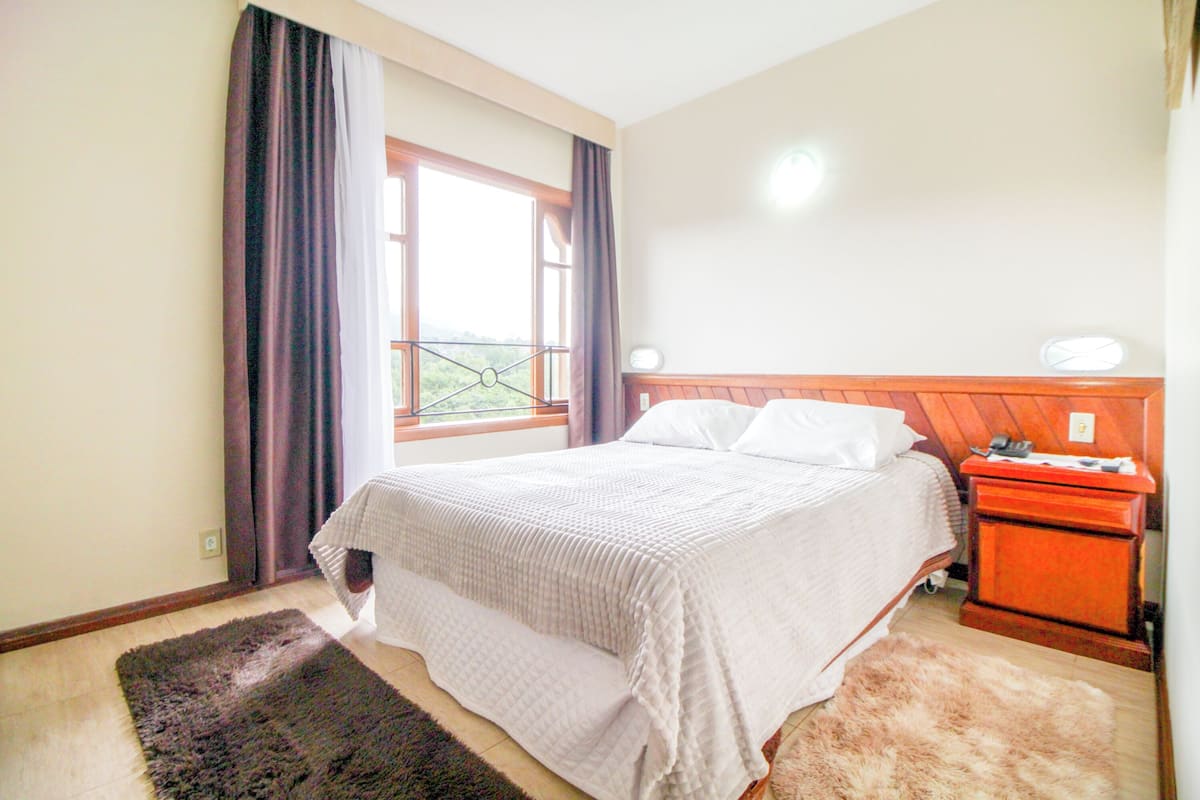 Une chambre à coucher avec un lit, une table de chevet, une fenêtre avec des rideaux et une vue sur la verdure à l’extérieur.