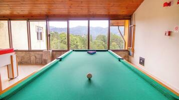 A pool table with a ball in the center, a red tablecloth, and a view of mountains through large windows.