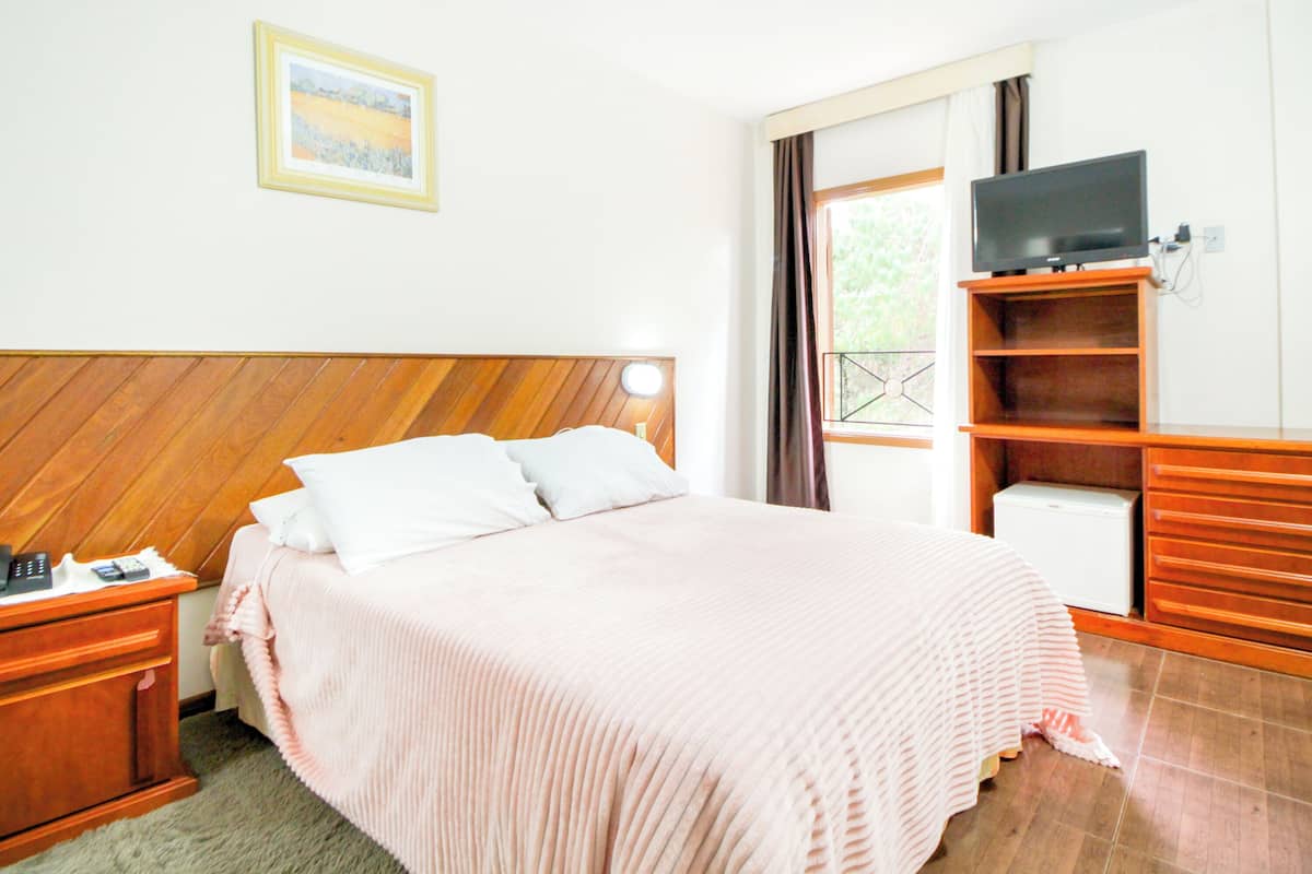 Une chambre à coucher avec une tête de lit en bois, un lit avec une literie blanche, une table de chevet avec une lampe, un téléviseur posé sur un meuble en bois et une fenêtre avec des rideaux.