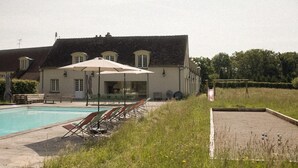 A swimming pool with lounge chairs and umbrellas, a covered seating area, and a building with a sloped roof in the background. - La Rambolitaine . (Poigny-la-Forêt)