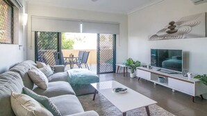A modern living room with a sofa, coffee table, TV, and a balcony with outdoor furniture. - ZEN Quest · ZEN Quest - 2bed/2bath Near Markets & Foreshore (Nightcliff)