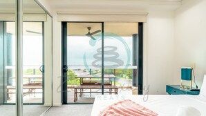 A hotel room with a bed, a nightstand, a desk lamp, and a sliding glass door leading to an outdoor area with a table and chairs. - Waterfront Retreat Ocean Views Pool Balcony (Darwin)