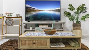 Een woonkamer met een grote flatscreen televisie, een houten salontafel, een rieten mand en een pot met een monstera plant.