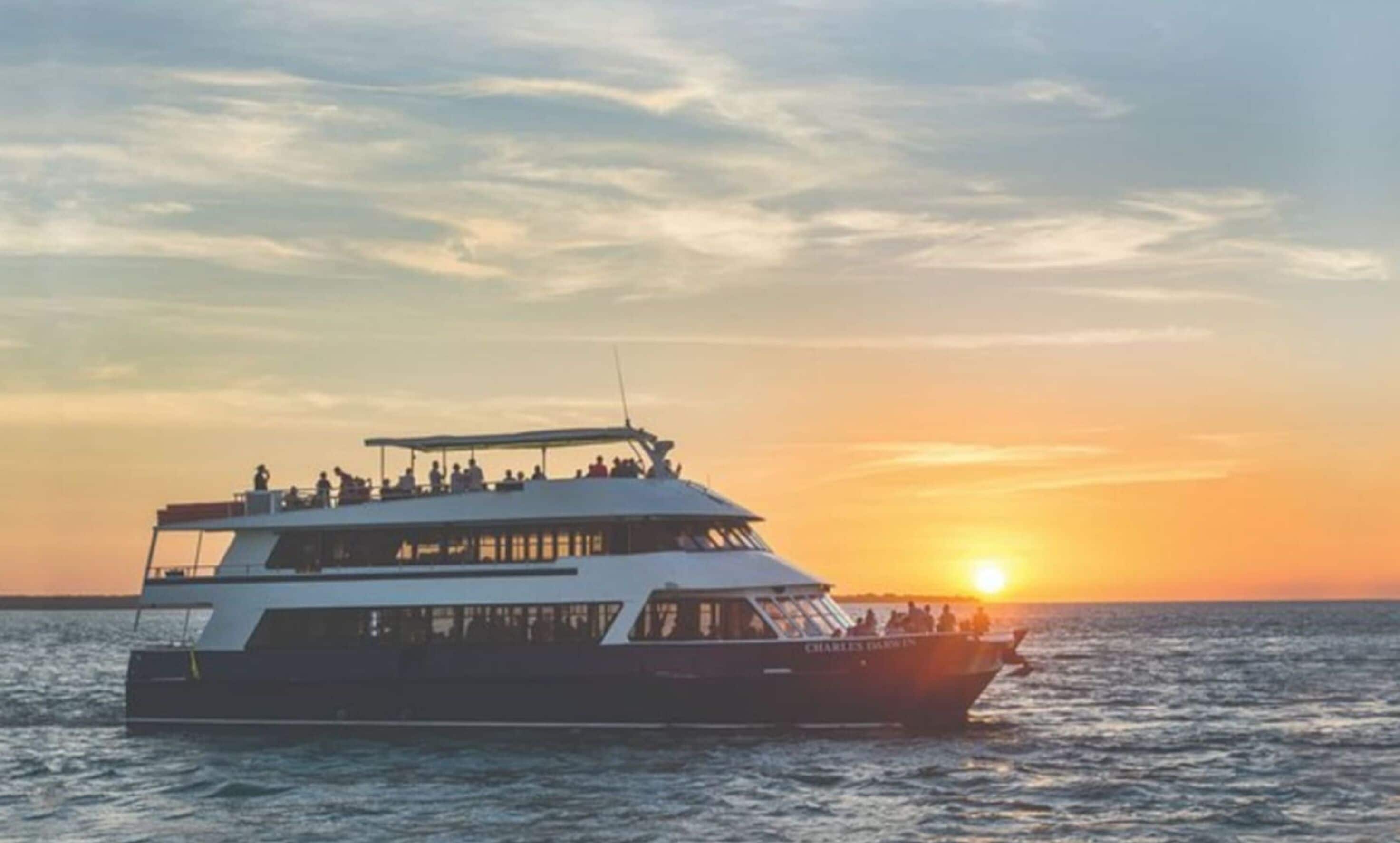 Ein großes, mehrstöckiges Boot mit dem Namen 'Charles Darwin' an der Seite, das bei Sonnenuntergang auf dem Wasser fährt.