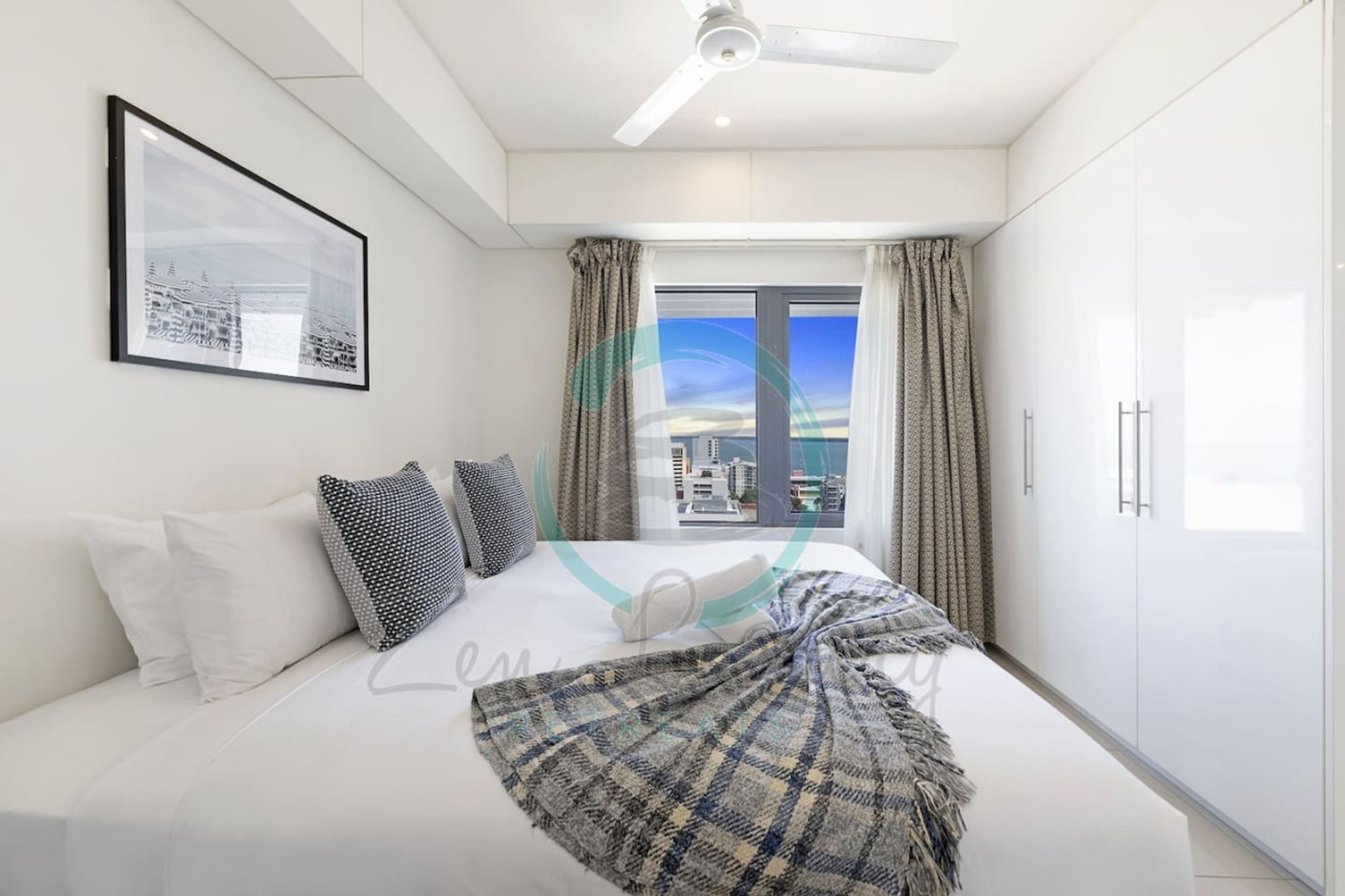 Une chambre d’hôtel moderne, équipée d’un lit, d’un ventilateur de plafond, d’un tableau encadré au mur et offrant une vue sur la ville par la fenêtre.