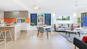 A modern kitchen with a central island, dining area with chairs, a grey sofa, and a coffee table. - Zen Nomad Balcony Outdoor Dining Markets (Nightcliff)