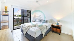 A modern bedroom with a bed, bedside table, and a view through a sliding glass door to the outside. - Zen Nomad Balcony Outdoor Dining Markets (Nightcliff)