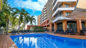 A swimming pool with lounge chairs and palm trees, surrounded by a multi-story hotel building with balconies. - Coastal Splendor Seaview Pool Balcony Dining (Darwin)