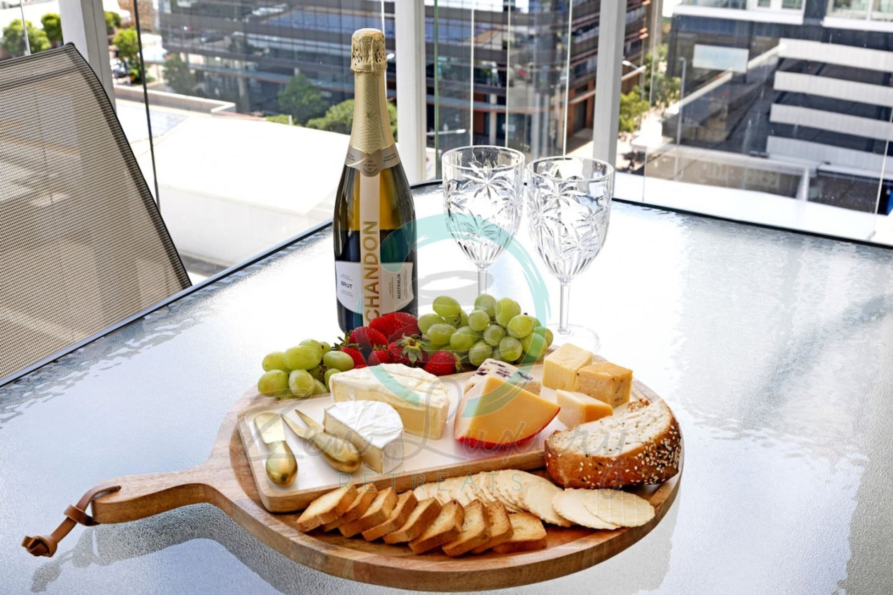 A glass table with a cheese board, grapes, strawberries, and a bottle of champagne.