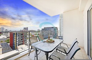 A balcony with a glass table and chairs, overlooking a cityscape with modern buildings. - Reef-side Splendor City Vistas Pool Workspace (Darwin)