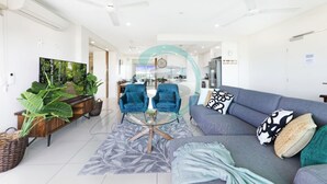 A modern living room with a sofa, two blue armchairs, a coffee table, and a large flat-screen TV. - Zen Seabreeze Workdesk Balcony Dining Seaview (Darwin)