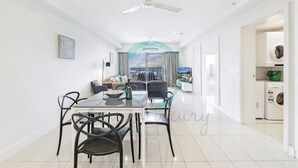 A modern living room with a dining area, a ceiling fan, and a view of the cityscape through large windows. - Zen Ocean Meadows Sea View - Pool - Outdoor Dining (Darwin)