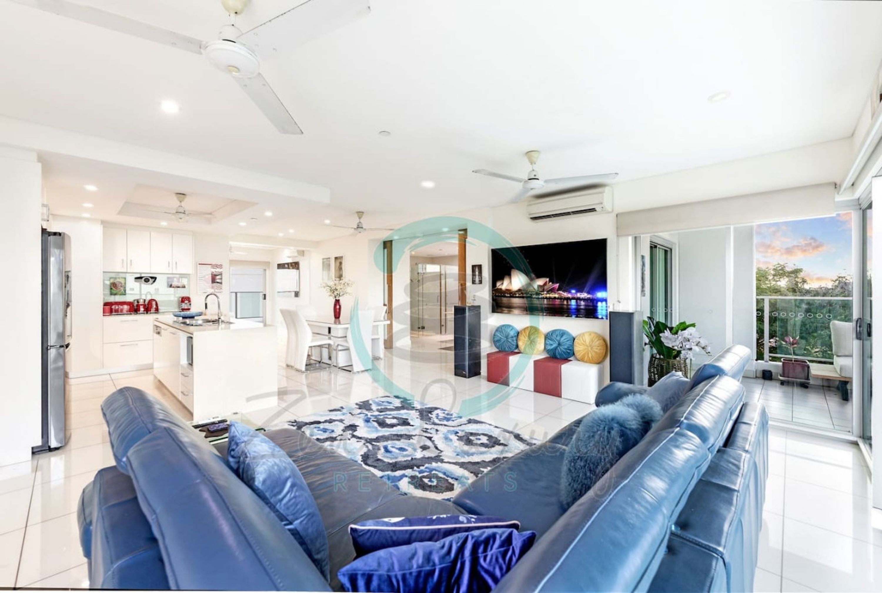 A modern living room with a sofa, a coffee table, and a TV. There is a kitchen area with cabinets and a refrigerator.