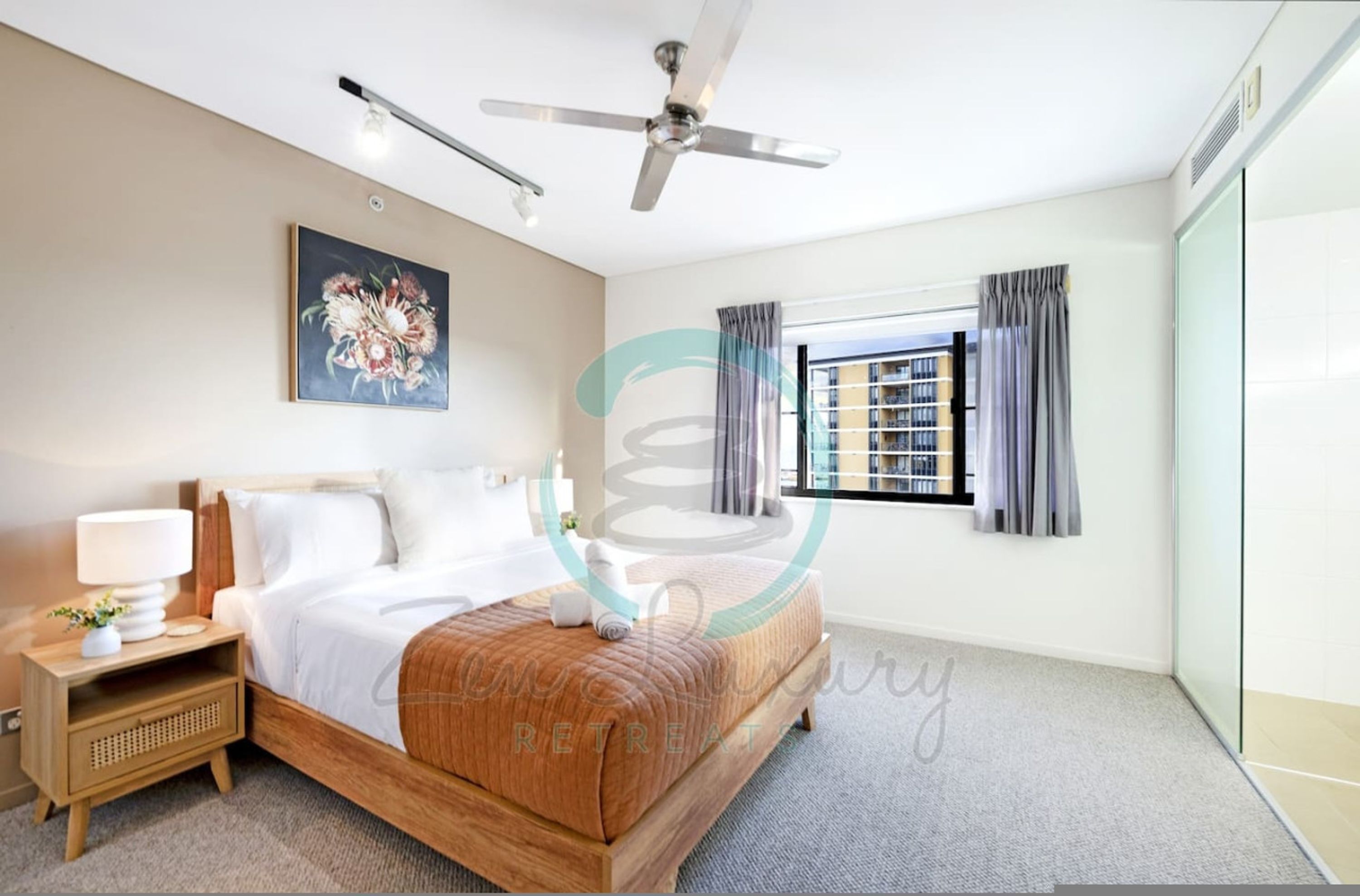 A modern hotel room with a bed, a nightstand, a ceiling fan, a window with curtains, and a painting on the wall.