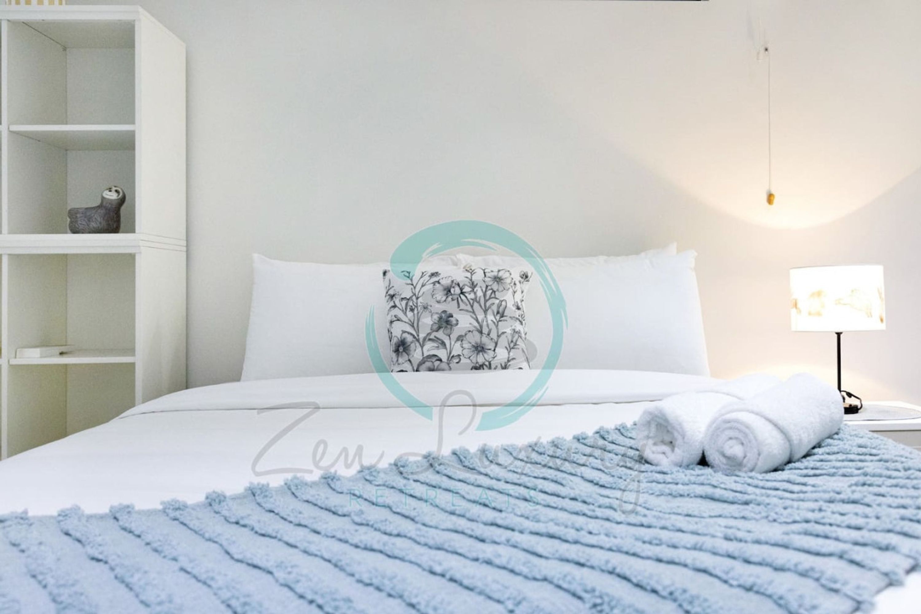 Un lit bien fait, agrémenté d’un coussin à motifs floraux, d’une couverture bleue texturée et de serviettes enroulées sur une table de chevet.