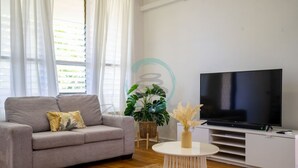 Ein Wohnzimmer mit grauem Sofa, einem Flachbildfernseher, einem kleinen weißen Tisch und einer Topfpflanze.