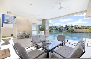 A modern outdoor dining area with a glass table and chairs, a grill, and a view of a lake. - Luxury Marina Mansion Pool Gym Bbq-office (Bayview)
