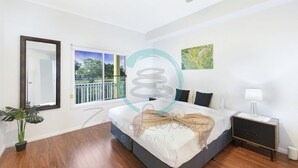 Un dormitorio con cama, mesita de noche, planta en maceta, ventana y una obra de arte en la pared.