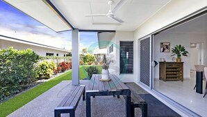 A modern patio with a table and benches, a ceiling fan, and a view of a garden and neighboring buildings. - Zenhouse Vista Elegant Coastal Haven (Muirhead)