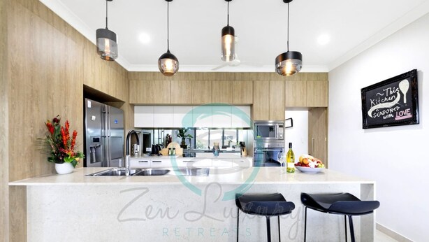 A modern kitchen with a central island, pendant lighting, and a sign that reads "This Kitchen Scored with Love". - Zenhouse Vista Elegant Coastal Haven (Muirhead)