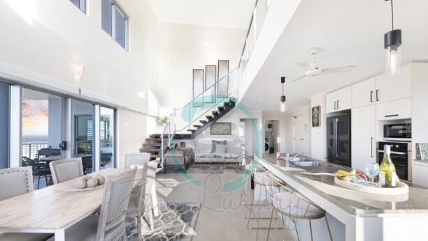 A modern kitchen with a central island, a dining area with a large table and chairs, and a staircase leading to an upper level. - Pinnacle Penthouse Ambience Views Pool Gym (Darwin)