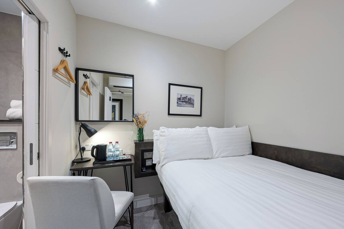 Une chambre d’hôtel avec un lit, un bureau et une chaise, un miroir, un tableau encadré au mur et un vase avec des fleurs.