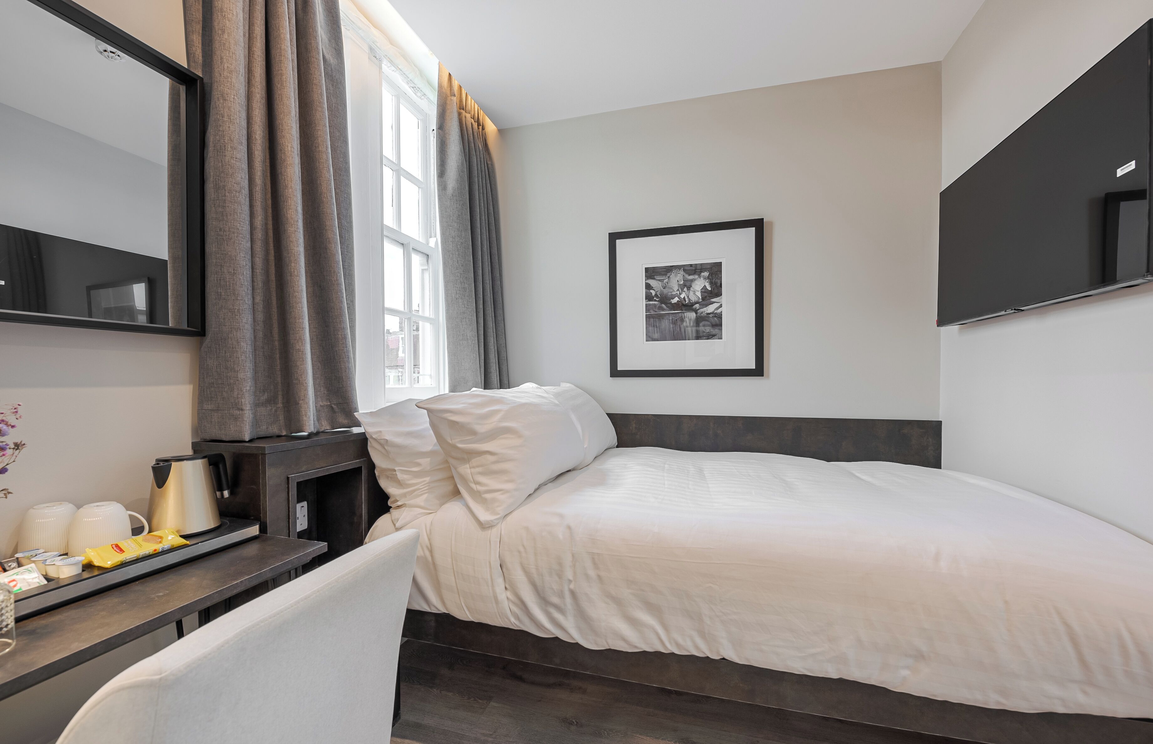 A hotel room with a bed, a bedside table with a kettle and cups, a framed picture on the wall, and a window with curtains.