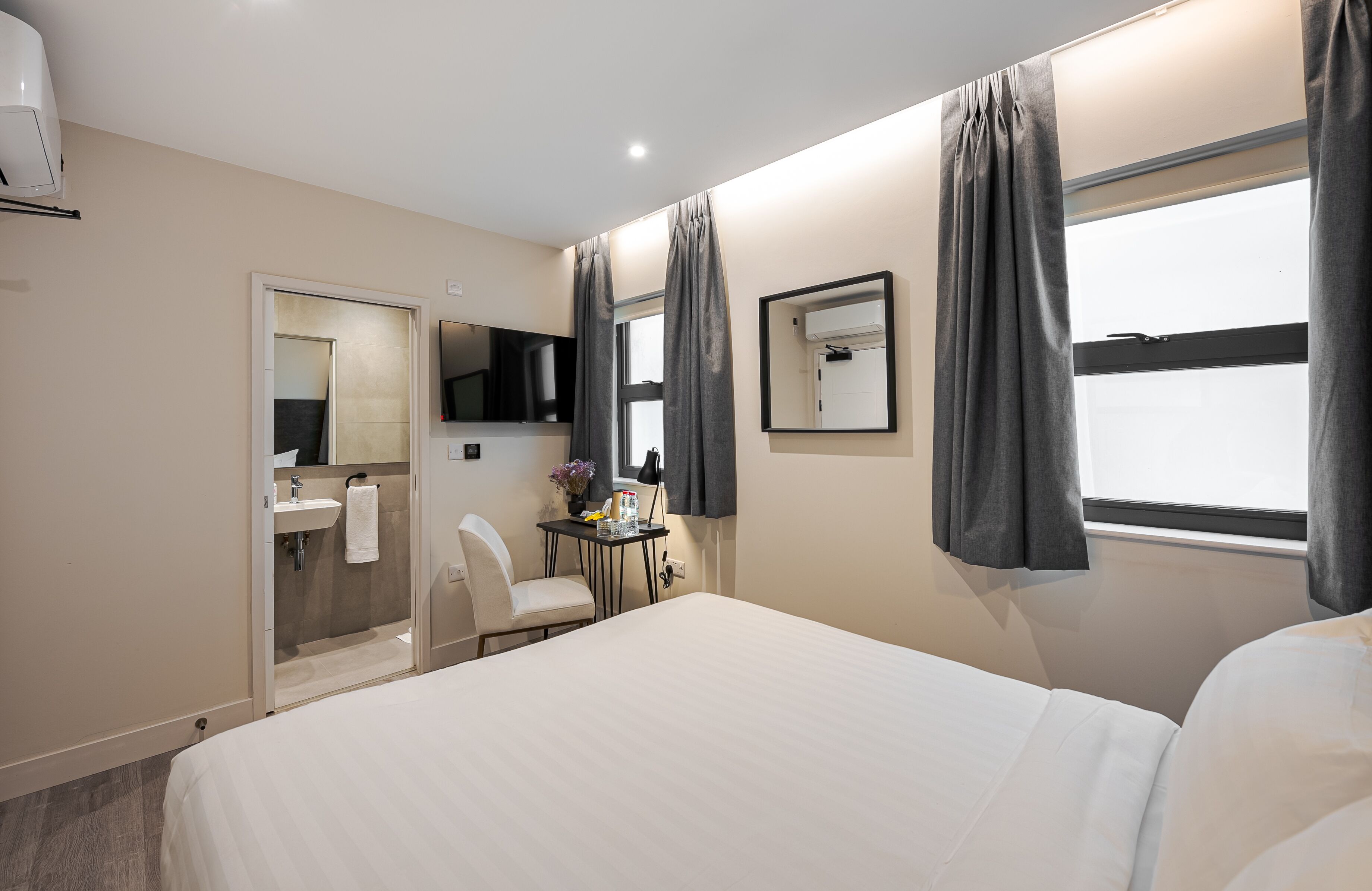 Una habitación de hotel con una cama, un escritorio y una silla, un espejo, una ventana con cortinas y un baño con lavamanos y ducha.