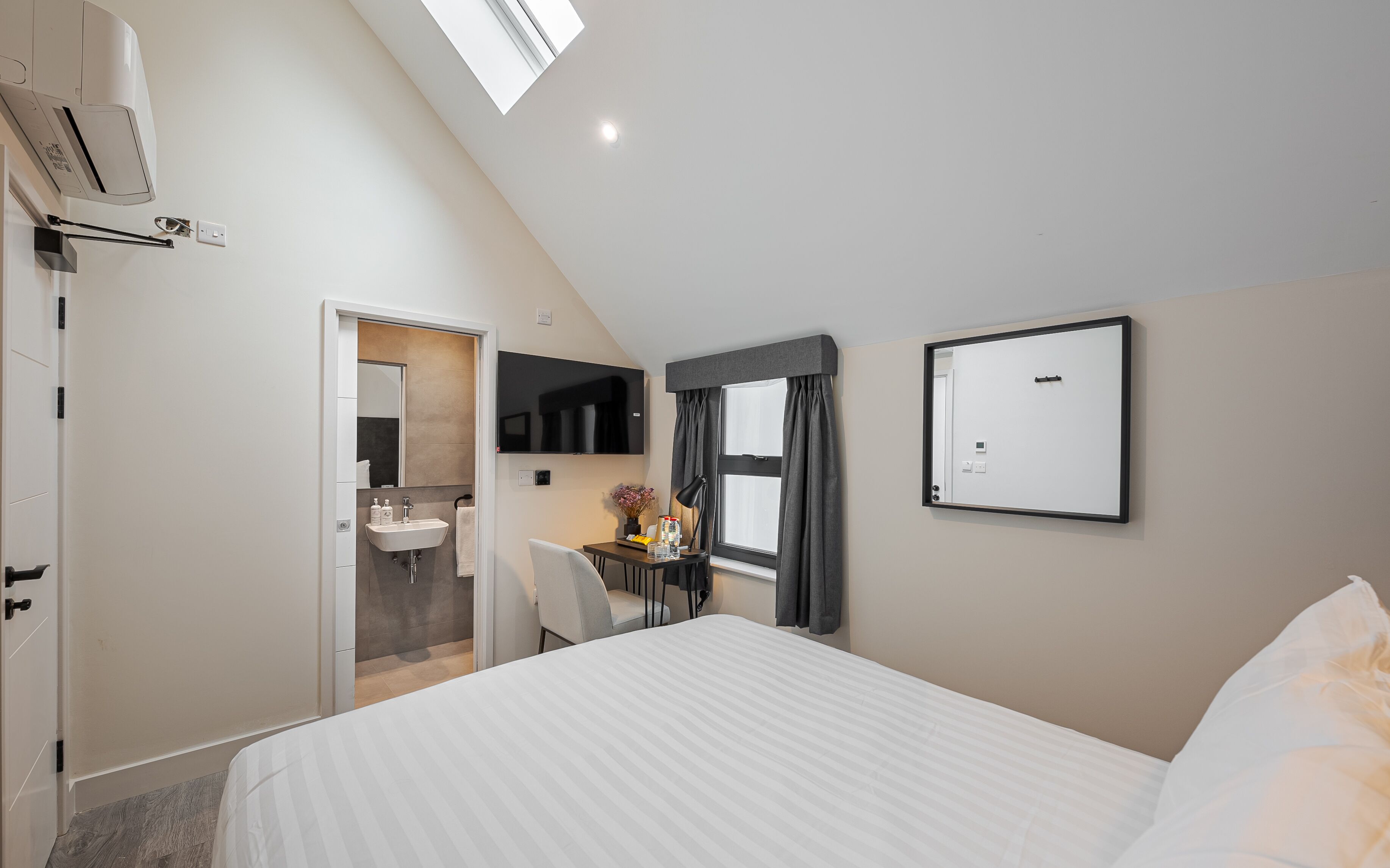 A modern hotel room with a bed, a desk with a chair, a bathroom with a sink, and a large mirror.