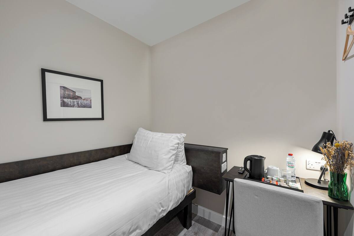 Une chambre d’hôtel bien rangée, avec un lit simple, une table de chevet avec une lampe, un petit bureau et un tableau encadré au mur.