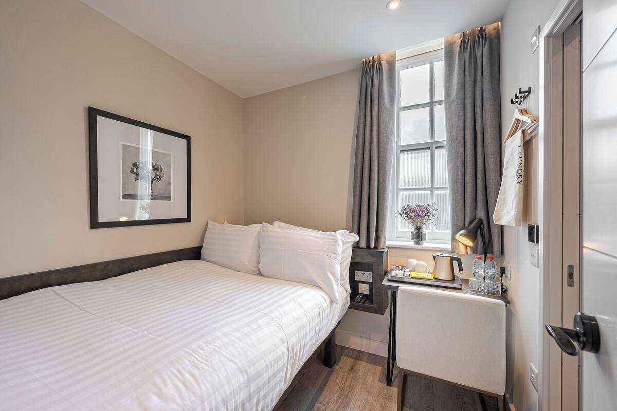 Une chambre d’hôtel avec un lit, une petite table sur laquelle se trouve une bouilloire, un tableau encadré au mur et une fenêtre avec des rideaux.