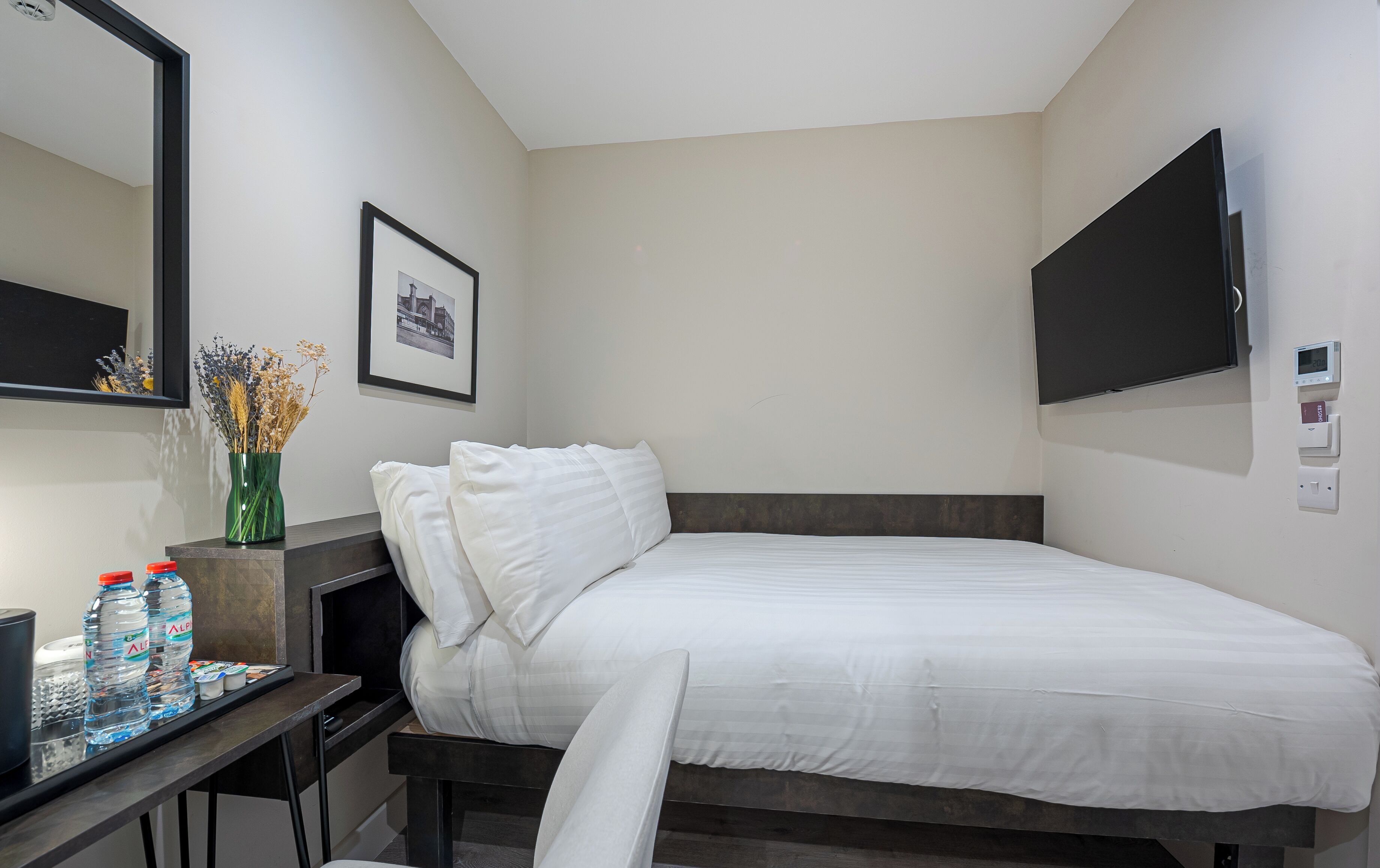 A hotel room with a bed, a desk with water bottles, a framed picture, a TV, and a thermostat.