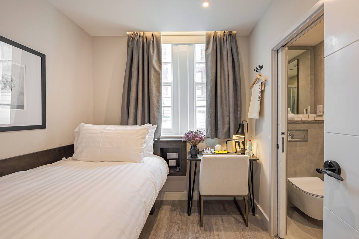 Une chambre d’hôtel avec un lit, un bureau, une chaise, une petite table ornée d’un vase de fleurs et une salle de bain visible à travers une porte ouverte.